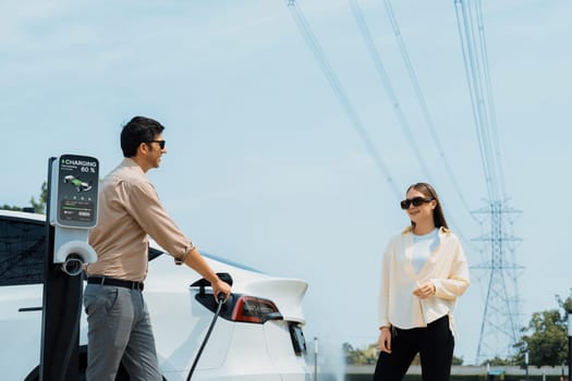 Young couple recharge EV car battery at charging station connected to power grid tower electrical industrial facility as electrical industry for eco friendly vehicle utilization. Expedient