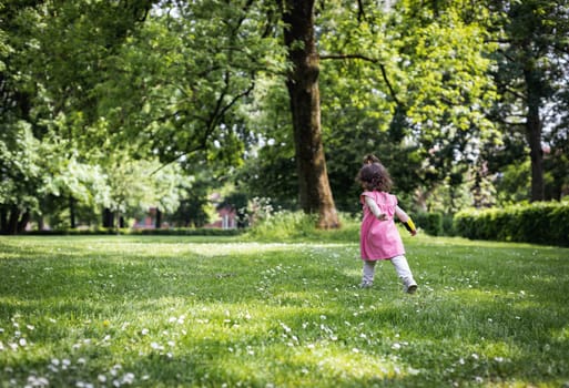 Portrait of one little baby girl in a pink dress running from the back through the city park at a picnic on a sunny summer day.