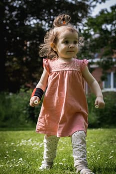 Portrait of one beautiful Caucasian girl with a ponytail on top of her head in a pink dress holds a meadow daisy in her hands and walks with an emotionally fast step along the lawn on a summer day in a public park, bottom side view close-up.