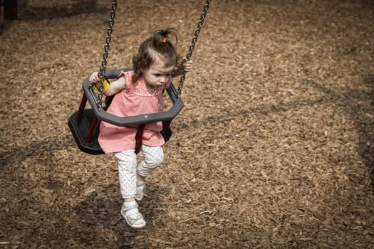 Portrait of one beautiful Caucasian baby girl in a pink dress and with a ponytail on the top of her head rides on a swing on a summer day in the park on the playground, top side view close-up.