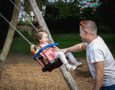 One handsome young Caucasian man father rides his little and happy laughing daughter girl on a swing on a summer day in the park on the playground, close-up side view.