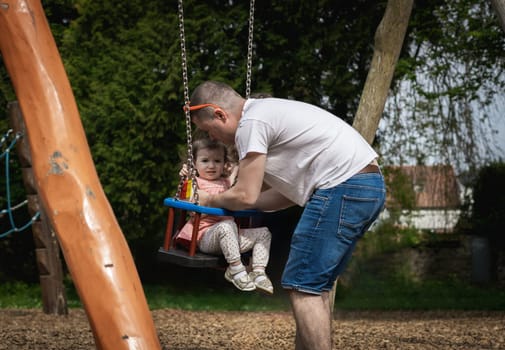 One handsome young Caucasian male father takes his little naughty baby girl daughter out of the swing on a summer day in the park on the playground, close-up side view.