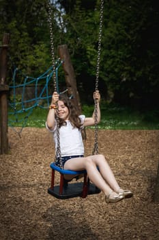 Portrait of one beautiful Caucasian brunette girl with a happy emotion and a smile on her face rides on a swing on a summer day in the park on the playground, close-up side view.