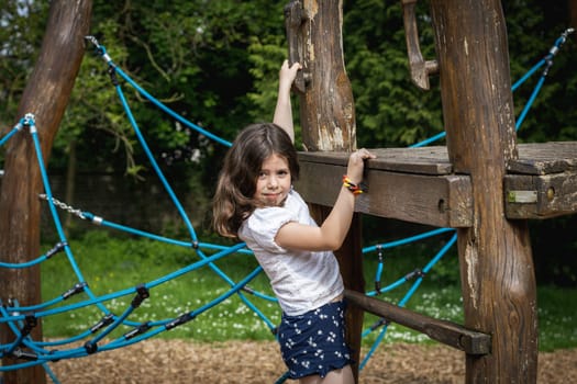 Portrait of one beautiful Caucasian brunette girl climbing on a wooden rope swing on a playground on a summer day in a park on a playground, close-up side view.