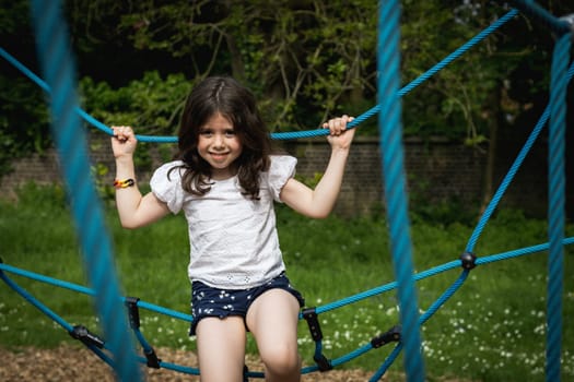 Portrait of one beautiful Caucasian happy brunette girl sitting holding hands on a blue rope swing on the playground on a summer day in the park at the playground, side view close-up.