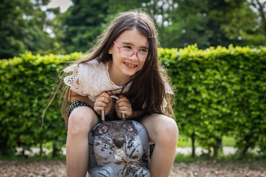 Portrait of one beautiful Caucasian happy brunette girl in glasses with long flowing hair swinging merrily on a wooden spring swing in the shape of a hare at the playground on a summer day in the park at the playground, side view close-up.