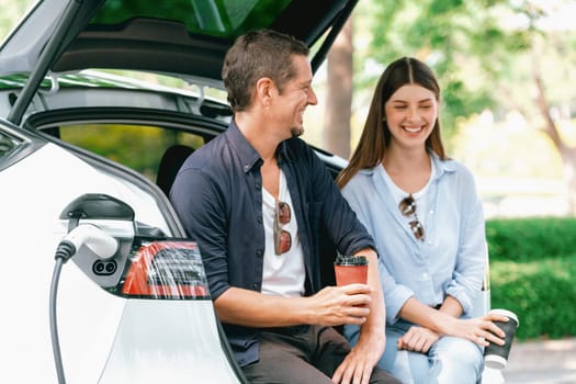 Lovely young couple drinking coffee while recharging battery for electric car during road trip travel EV car in natural forest or national park. Eco friendly travel during vacation and holiday. Exalt