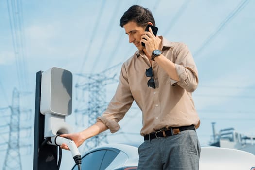 Man talking on the phone while recharge EV car battery at charging station connected to power grid tower electrical as electrical industry for eco friendly car utilization.Expedient