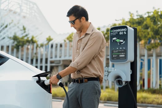 Young man put EV charger to recharge electric car's battery from charging station in city commercial parking lot. Rechargeable EV car for sustainable environmental friendly urban travel. Expedient