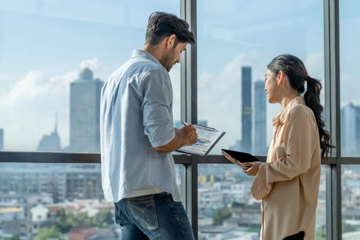 Professional businessman present financial statistic to investor while standing near panoramic window shows skyscraper or city view. Diverse businesspeople discuss about marketing plan. Tracery.