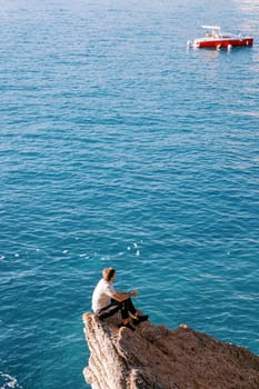 Young man sits on a rock ledge and looks at a boat sailing on the sea. Back view. High quality photo