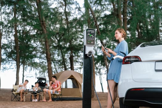 Outdoor adventure and family vacation camping at sea travel by eco friendly car. Woman or mother check car's battery with smartphone while charging EV car frin charging station in campsite. Perpetual