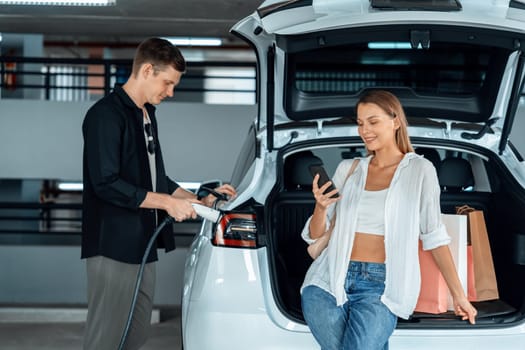 Young couple travel with EV electric car to shopping center parking lot charging in downtown city showing urban sustainability lifestyle by green clean rechargeable energy of electric vehicle innards