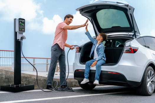 Family road trip vacation traveling by the sea with electric car, father and son high five after reach destination at EV charging station by the seashore. Eco-friendly car for environment. Perpetual