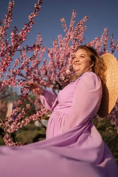 Woman blooming peach orchard. Against the backdrop of a picturesque peach orchard, a woman in a long pink dress and hat enjoys a peaceful walk in the park, surrounded by the beauty of nature