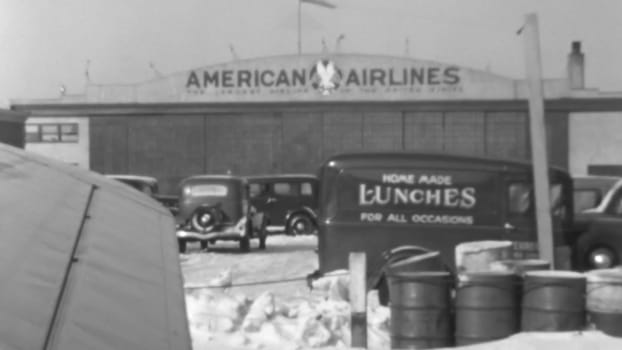 Unique perspective from inside a 1930s airliner. Photo shows airport hangars, other structures, and fuel drums on the tarmac, captured through the airplane window. Boeing 247 of United Air Lines