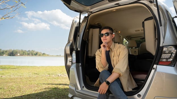 Carefree young man in sunglasses sitting in the open trunk of minivan near lake with mountains and clear sky.