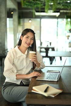 Smiling millennial female worker holding paper cup and working with laptop at coffee shop.