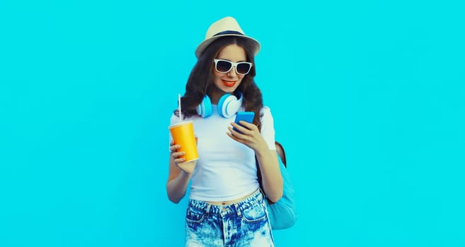 Portrait of modern caucasian young woman listening to music in headphones with mobile phone holding cup of fresh juice on blue studio background