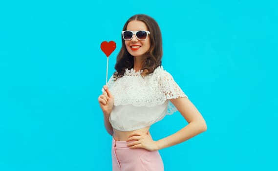 Portrait of beautiful young woman with red heart shaped lollipop on stick wearing white sunglasses on blue studio background