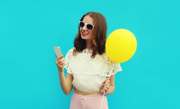 Portrait of happy smiling caucasian young woman 20s with mobile phone looking at device holding colorful yellow balloon on studio blue background