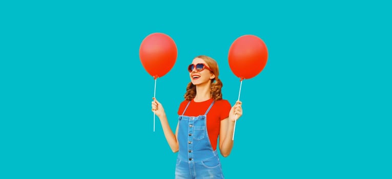 Happy cheerful young woman having fun with balloon wearing red heart shaped sunglasses, jumpsuit on blue background