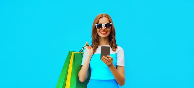 Beautiful happy young woman looking at mobile phone with shopping bag wearing summer dress on blue studio background