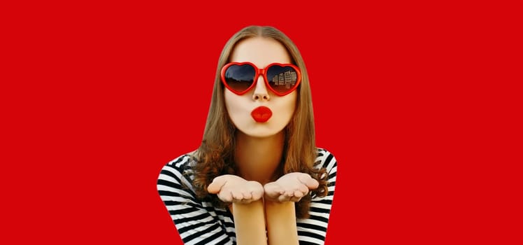 Portrait of beautiful young woman blowing her lips with red lipstick sends sweet air kiss wearing heart shaped sunglasses on studio background