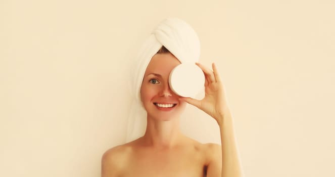 Natural beauty portrait of happy smiling young caucasian woman touches her clean skin applying face cream while drying wet hair with white wrapped bath towel on her head
