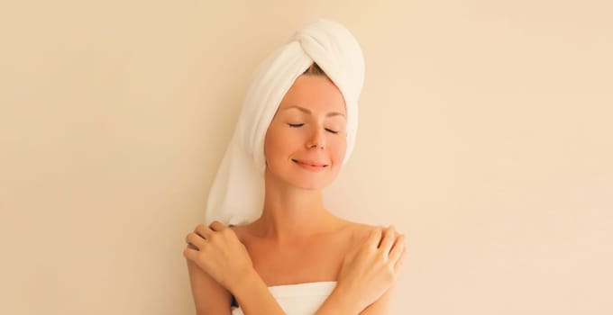 Natural beauty portrait of happy smiling young caucasian woman touches her clean skin while drying wet hair with white wrapped bath towel on her head