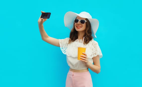 Summer portrait of beautiful happy smiling young woman taking selfie with mobile phone and cup of fresh juice wearing white straw hat on colorful blue background