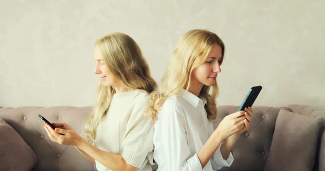 Two caucasian women with mobile phone looking at device, middle aged mother or sister and adult daughter together sitting on couch in living room at home