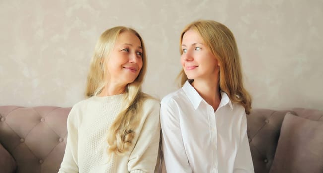 Portrait of happy smiling caucasian middle aged mother and adult daughter looking at each other together sitting on couch at home