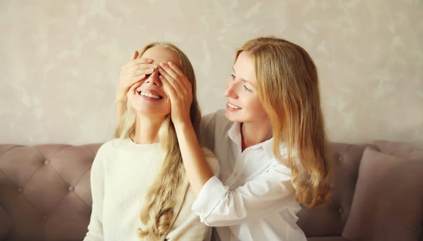 Happy smiling caucasian adult daughter covering mother's eyes making surprise sitting on couch at home