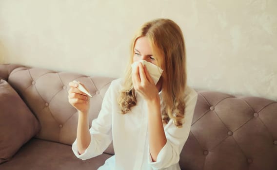 Sick exhausted woman sneezing blow nose using tissue and measuring checking body temperature looking at thermometer sitting on couch at home