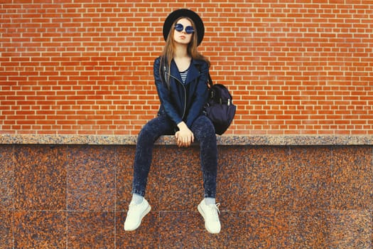 Portrait of stylish young woman model wearing black round hat, leather jacket in rock style on city street on brick wall background