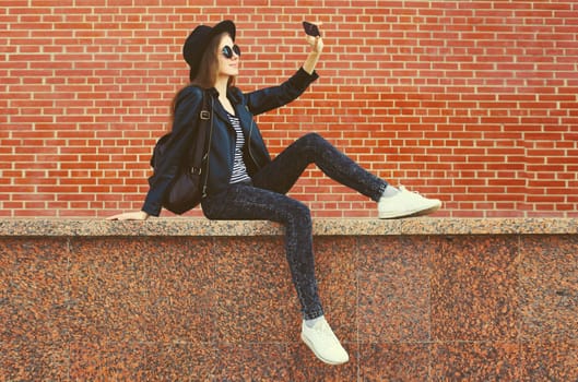 Stylish modern caucasian young woman taking selfie with mobile phone wearing black round hat, jacket and backpack on city street background