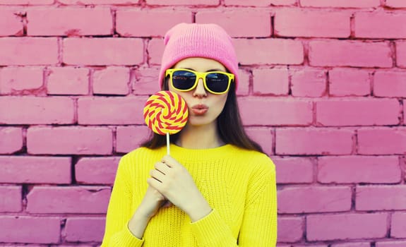 Colorful portrait of stylish modern young woman with lollipop blowing her lips sending sweet air kiss wearing vivid yellow sweater, sunglasses and pink hat on brick wall background
