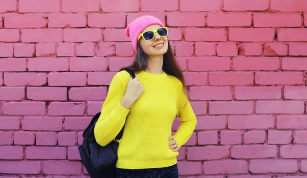 Portrait of stylish young teenager girl with backpack posing in vivid colorful clothes, knitted yellow sweater and pink hat, sunglasses on city street brick wall background