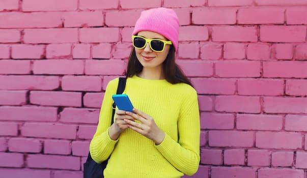 Modern caucasian young woman 20s model with mobile phone looking at device wearing colorful vivid clothes, yellow sweater and pink hat on city street