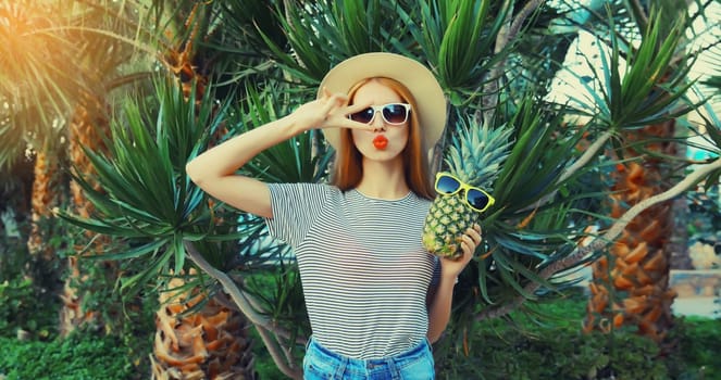Summer portrait of beautiful young woman with pineapple fruits posing blowing her lips sends kiss wearing sunglasses, straw hat on palm tree background
