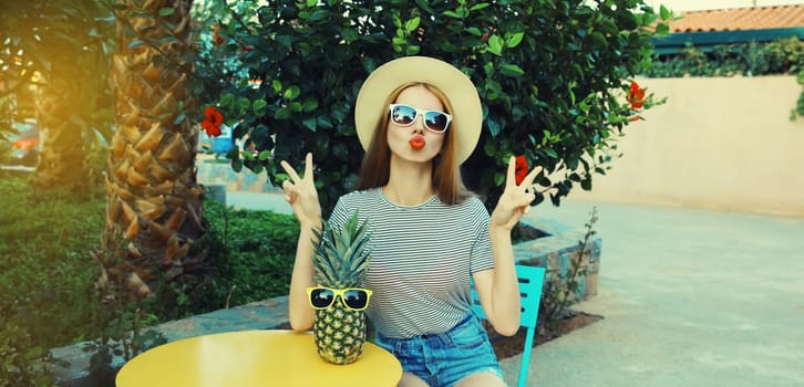 Summer portrait of beautiful young woman with pineapple fruits posing blowing her lips sends kiss wearing sunglasses sitting at the table in street cafe in city park