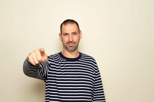 Portrait of handsome man in his 40s pointing index finger at you over isolated beige background, looking at camera. A friendly, smiling guy pointing his index finger at the camera choosing you.