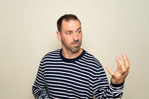 Bearded hispanic man in his 40s wearing a sweater looking extremely angry showing his claw hand while having an aggressive and defiant expression, isolated over beige background