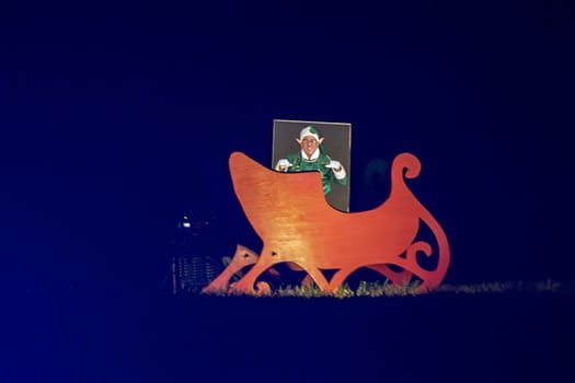 Nighttime View Of A Large Wooden Cut-Out Of Santa's Sleigh With A Person Posing As An Elf Through A Face-In-Hole Board, Creating A Festive Photo Opportunity.