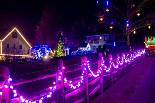 Night Scene Of A Street With Buildings And Trees Decorated With Christmas Lights And A Wooden Fence Lined With Purple Lights, Under A Sky With Star-Shaped Lights.