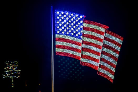 Nighttime Display Of Three American Flags Made Of Bright Lights Mounted On A Pole, With A Christmas Tree Light Display Visible To The Left Side.