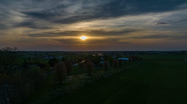 Dramatic Sunset Casting A Warm Glow Over A Rural Landscape With Silhouetted Trees And Buildings Under A Cloud-Streaked Sky.