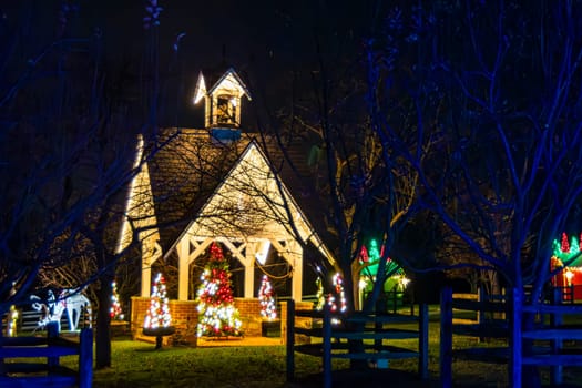 Night View Of A Small Chapel Decorated With Christmas Lights And Multiple Christmas Trees, Framed By Bare Trees And A Wooden Fence In A Serene Setting.