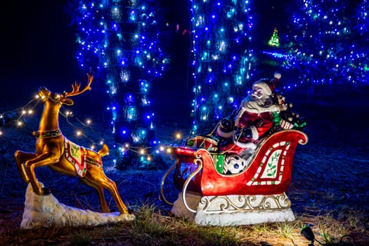 Colorful Nighttime Holiday Display Featuring A Lit Reindeer And Santa Claus In A Sleigh Decoration, Surrounded By Blue-Lit Trees And Festive Ornaments.